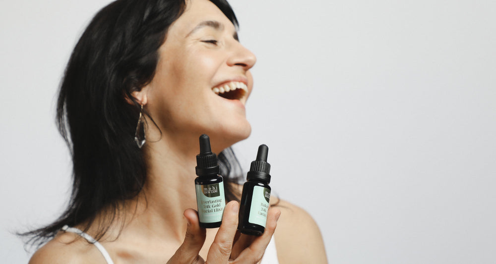 Model is laughing holding two bottles of 24K-Gold-face-oil by Mohi Skin Care