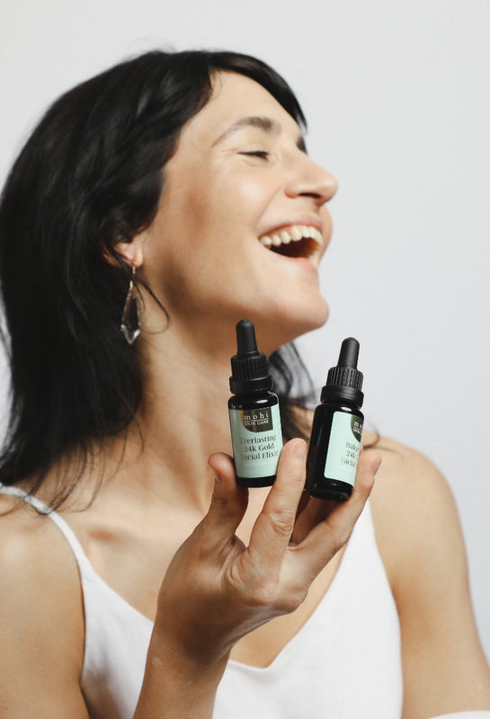Model is laughing holding two bottles of 24K-Gold-face-oil by Mohi Skin Care